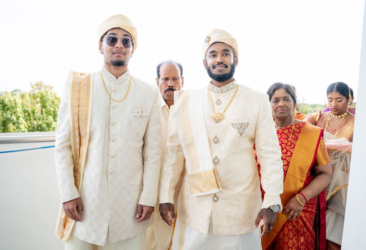 Two grooms in matching white and gold traditional Indian wedding attire pose with family at their wedding.