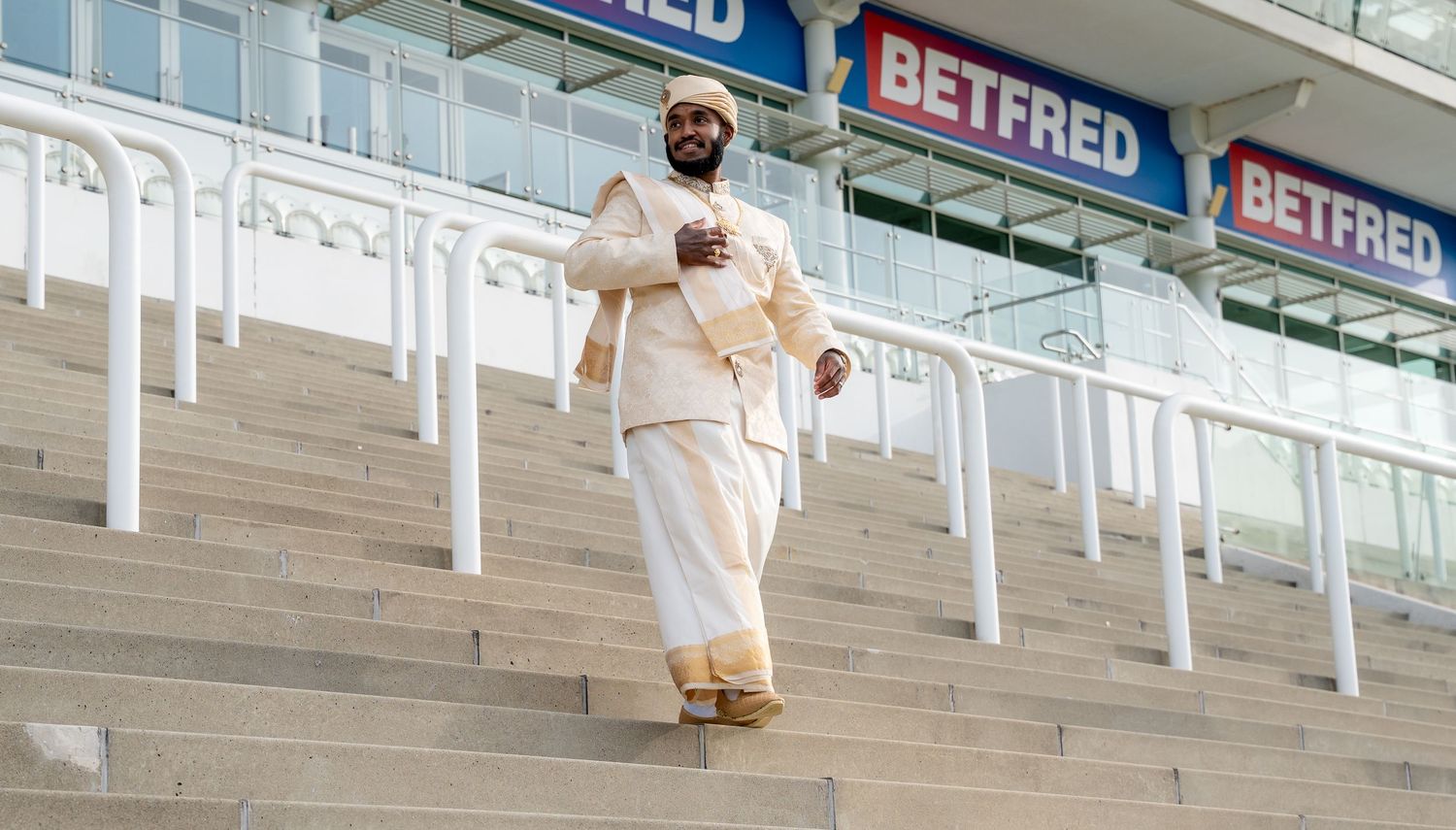 Man in white traditional South Asian attire walking down stadium steps near Betfred signage.
