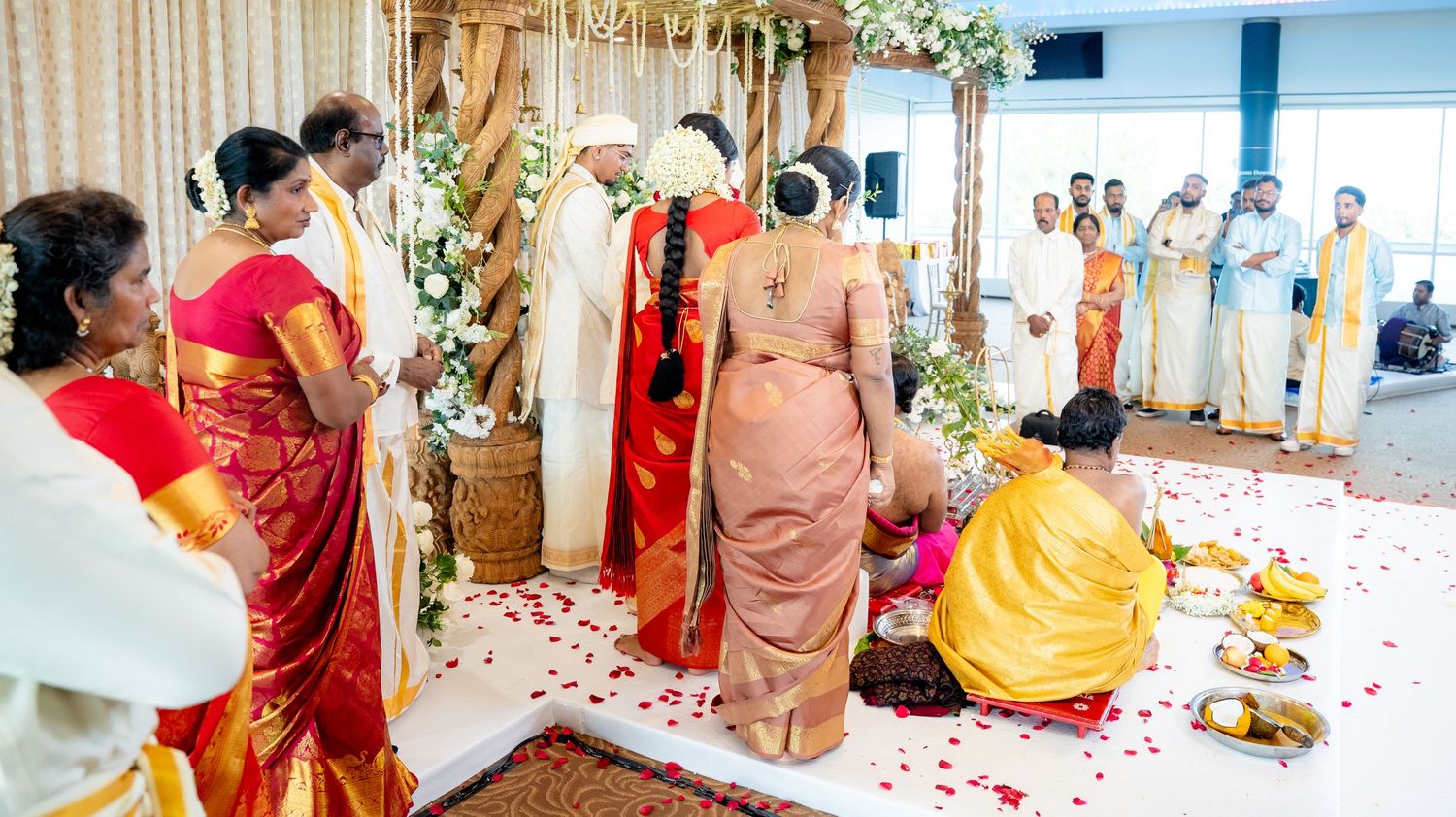 Family and guests gather around couple during traditional Hindu wedding ceremony on petal-strewn aisle.