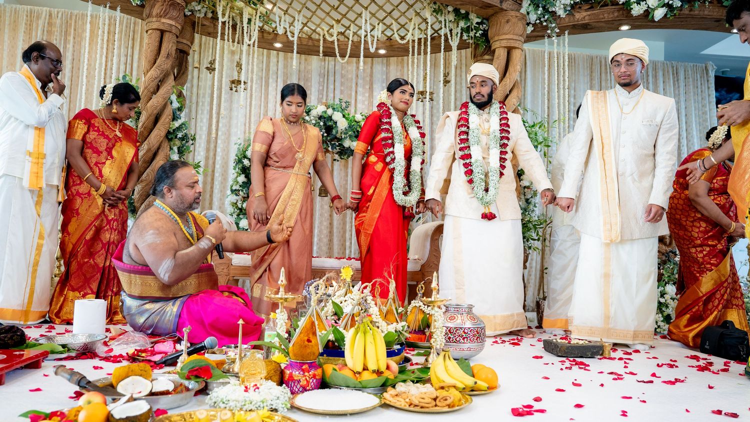 South Indian Hindu wedding ceremony with priest, family members, bride in red saree and groom in white on floral mandap.