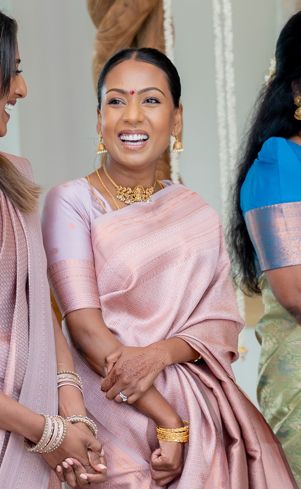 Woman in pink saree with gold jewelry smiling at an event, wearing traditional Indian attire and accessories.