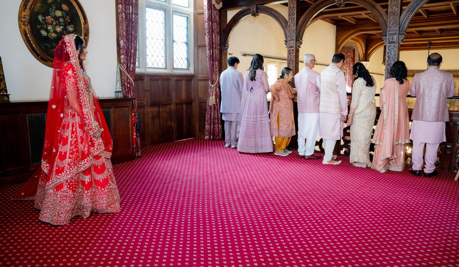Indian bride in red lehenga standing apart from wedding guests gathered in ornate hall.