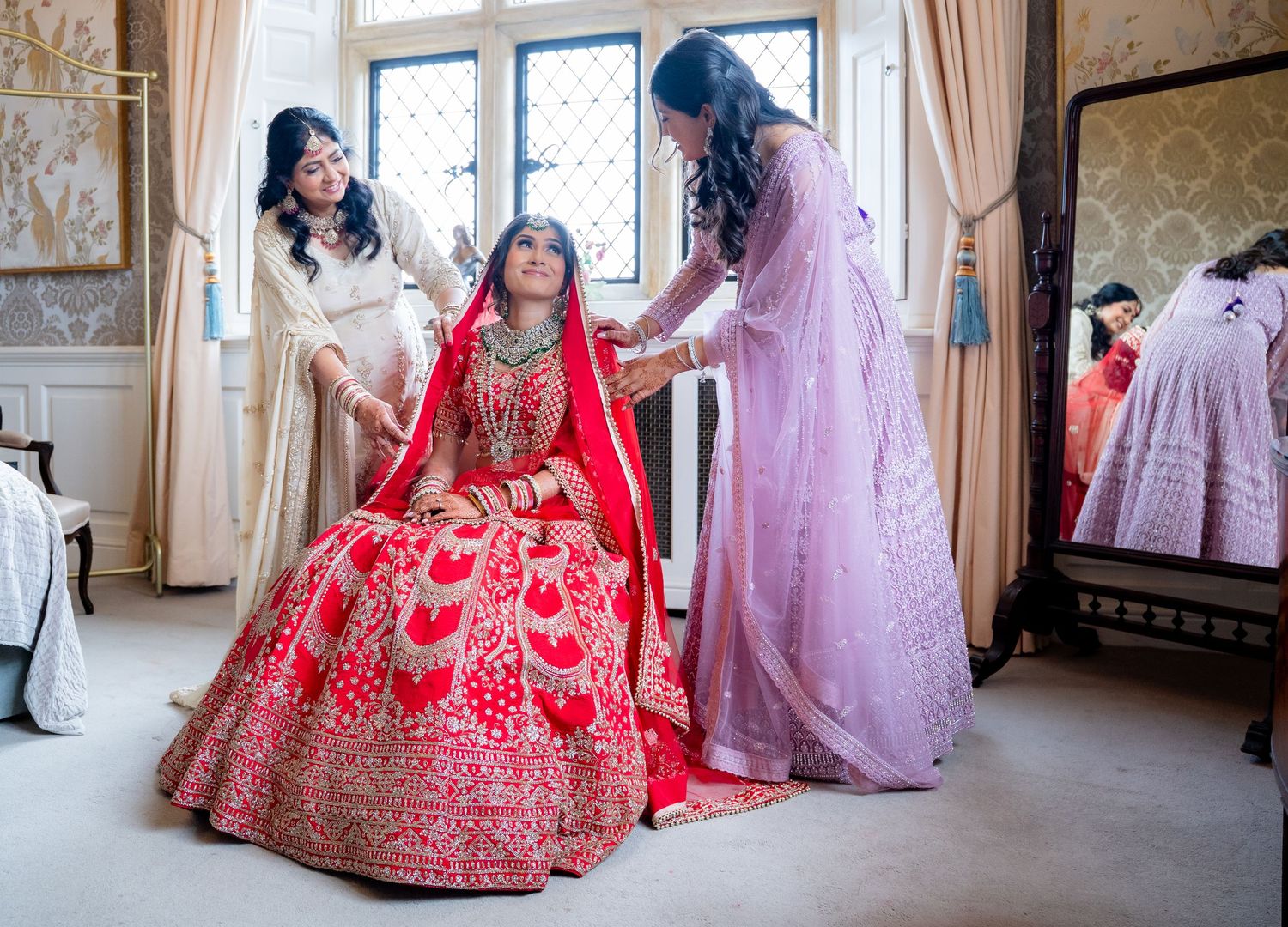 Two women help Indian bride in red lehenga with final preparations in an elegant room.