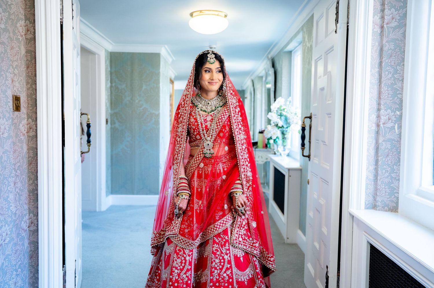 Indian bride in red lehenga with gold embroidery standing in elegant hallway.