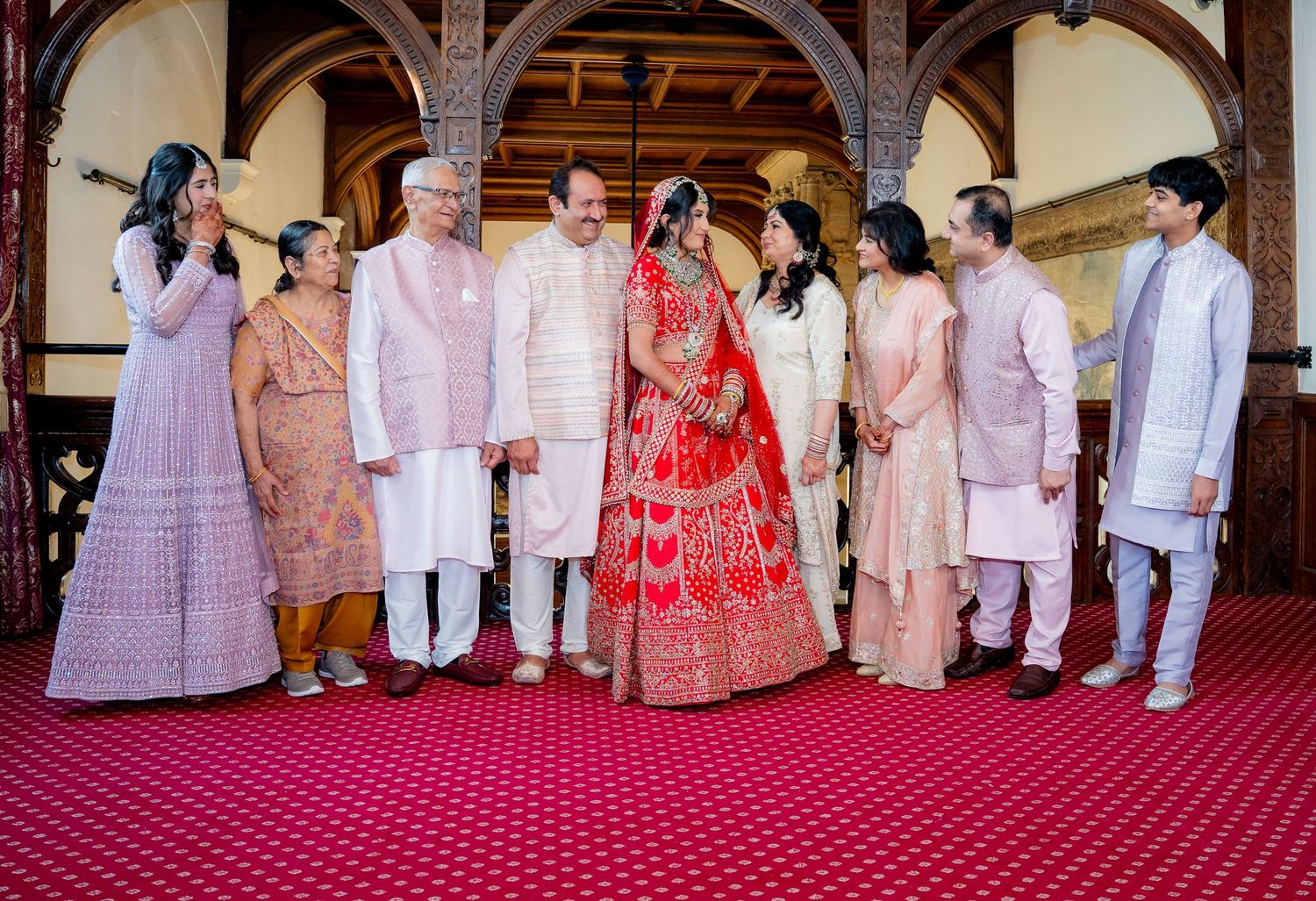 Indian wedding group photo with bride in red lehenga surrounded by family in elegant hall.