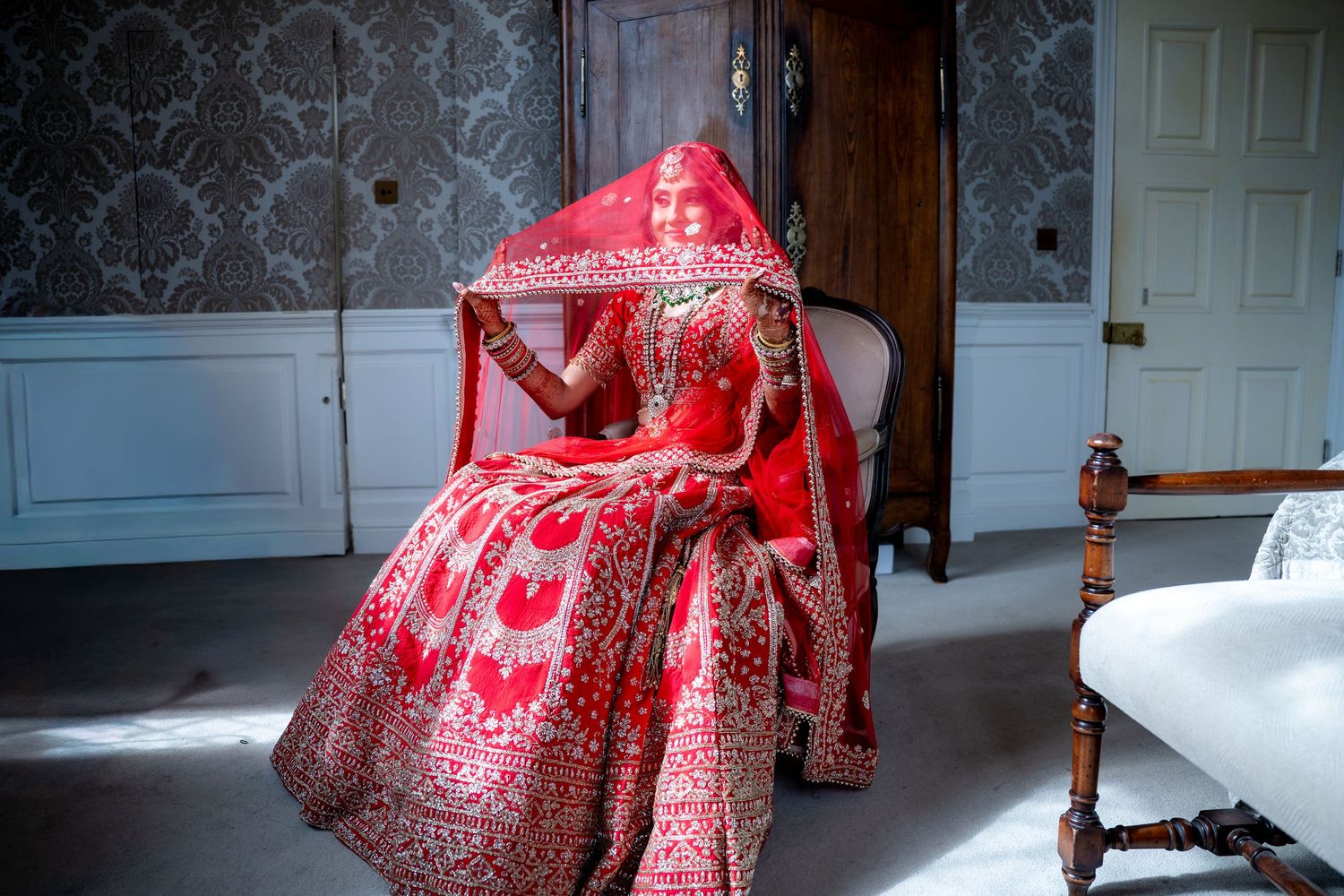 Indian bride in red embroidered lehenga and dupatta seated in sunlit ornate room with veil.