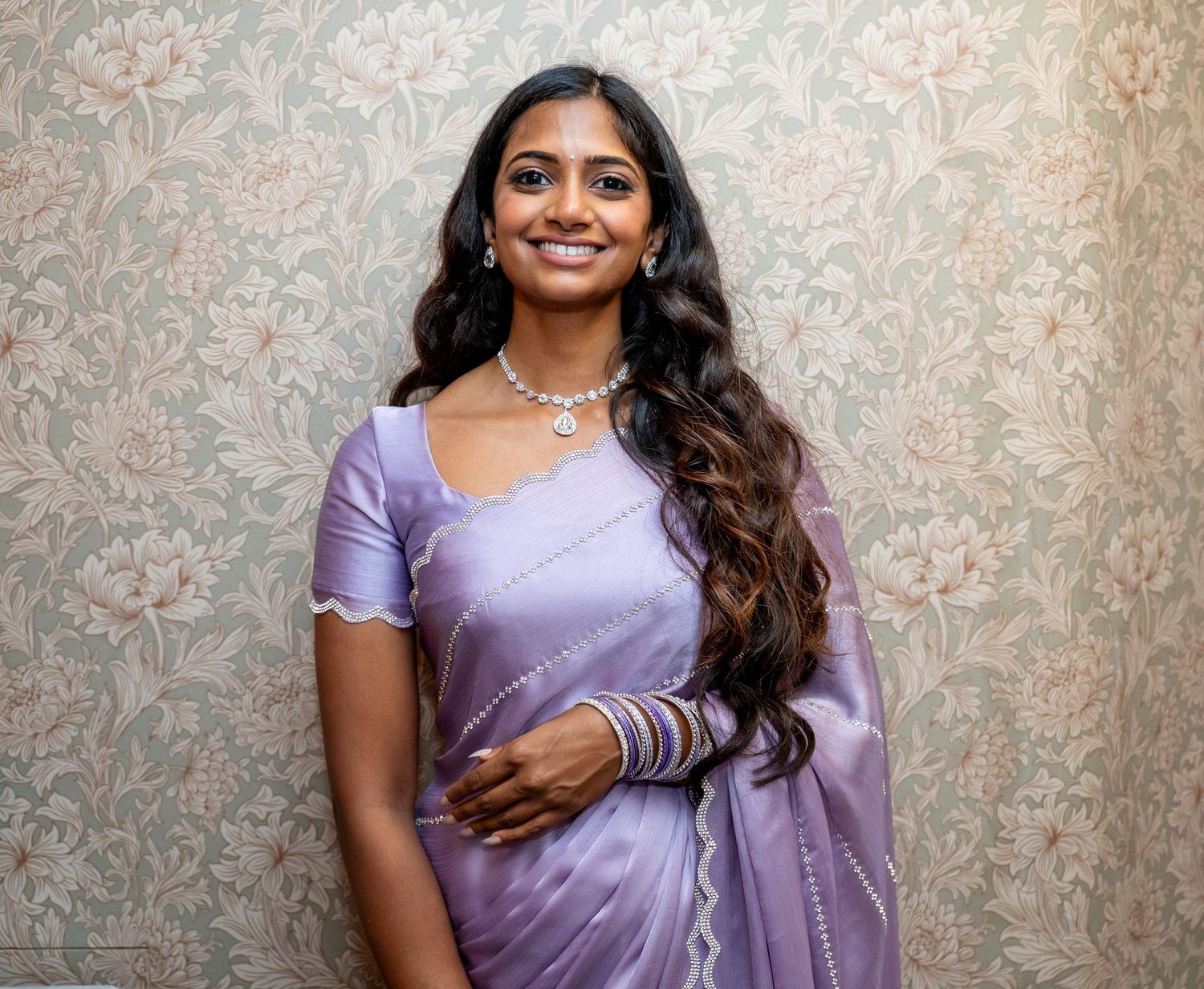 Woman in elegant purple saree with long wavy hair smiling at camera against decorative wallpaper background.