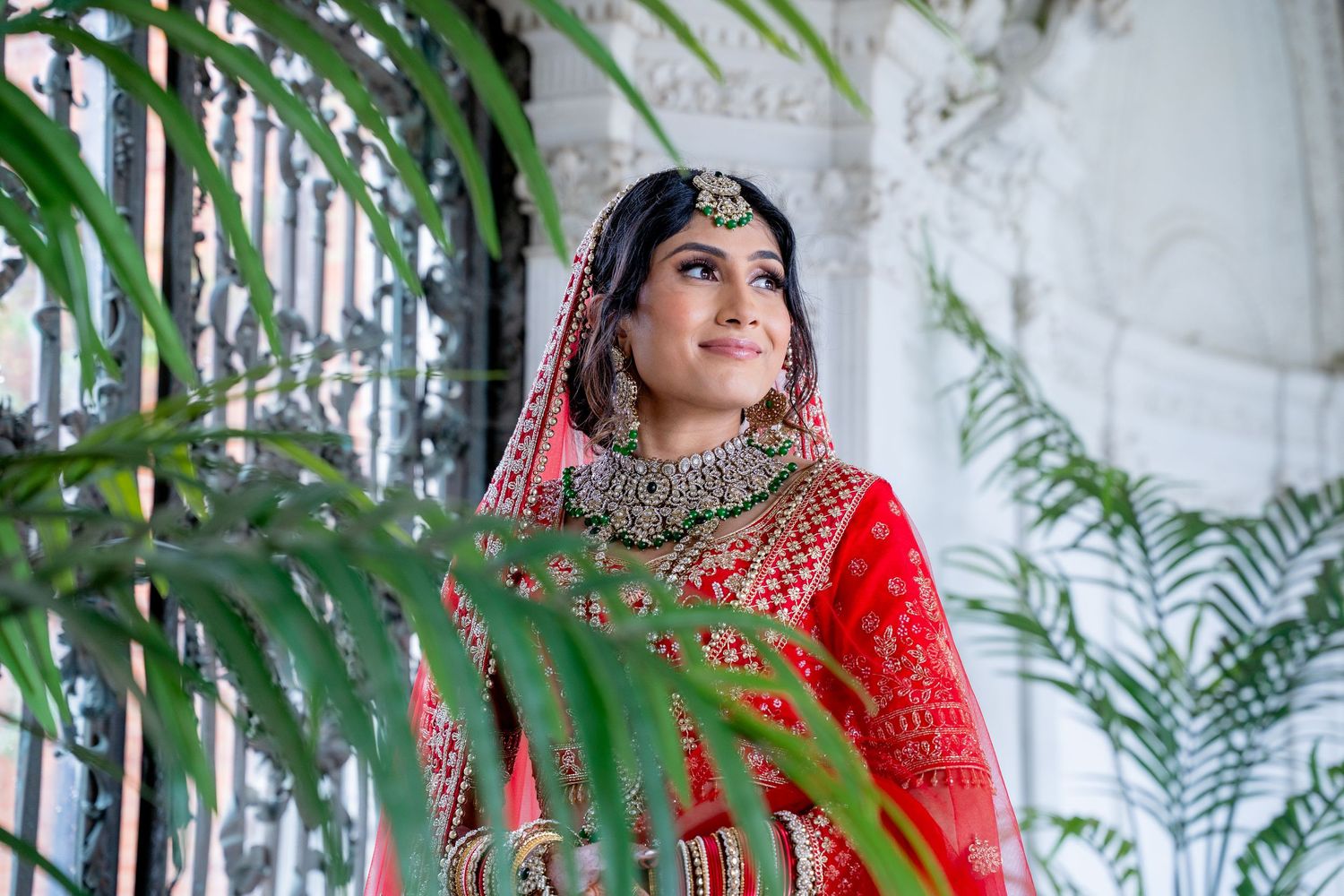 Beautiful South Asian bride in red bridal lehenga and gold jewelry gazes upward outdoors.