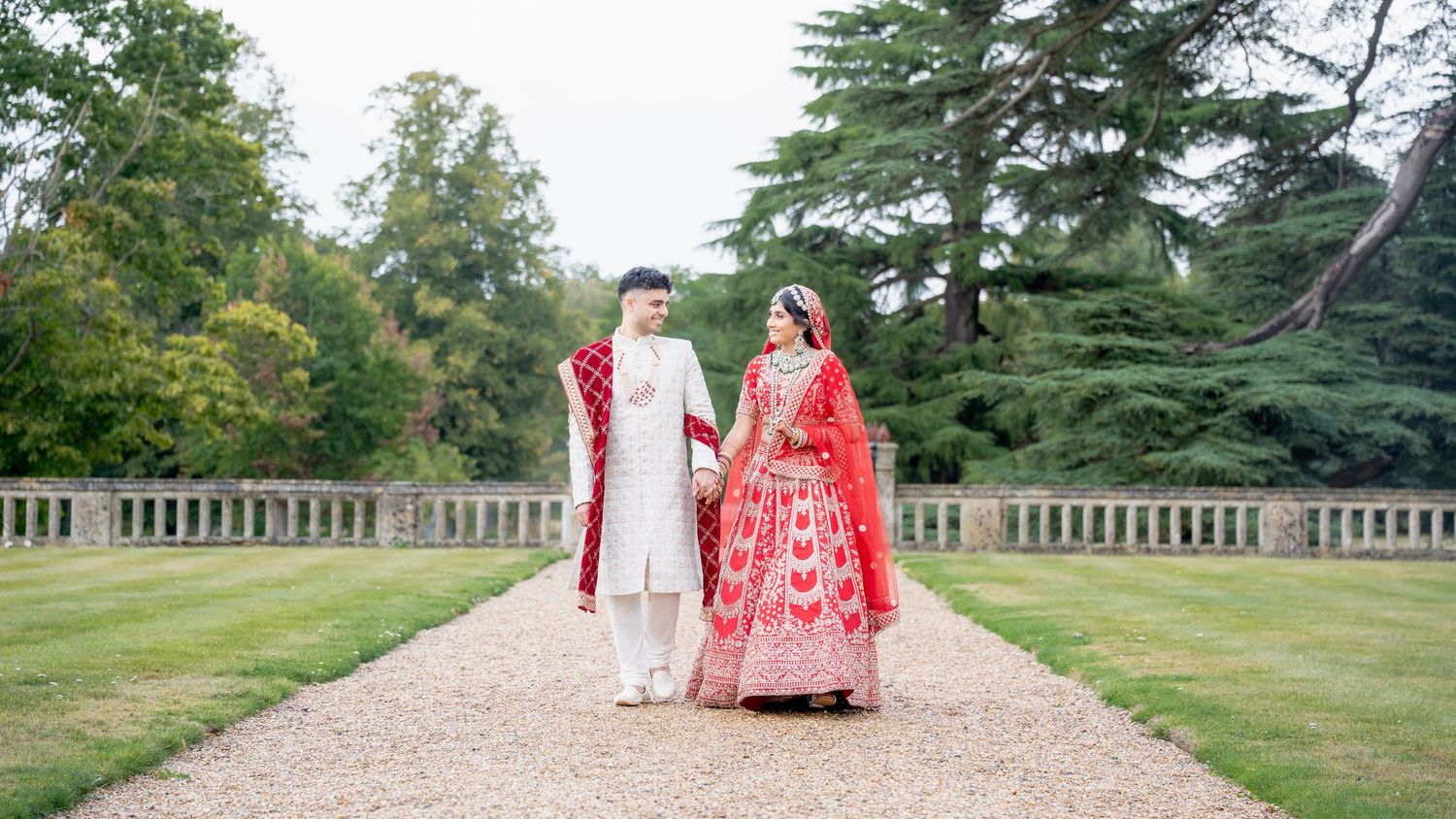 Indian wedding couple in traditional red and white attire walking hand in hand in garden.