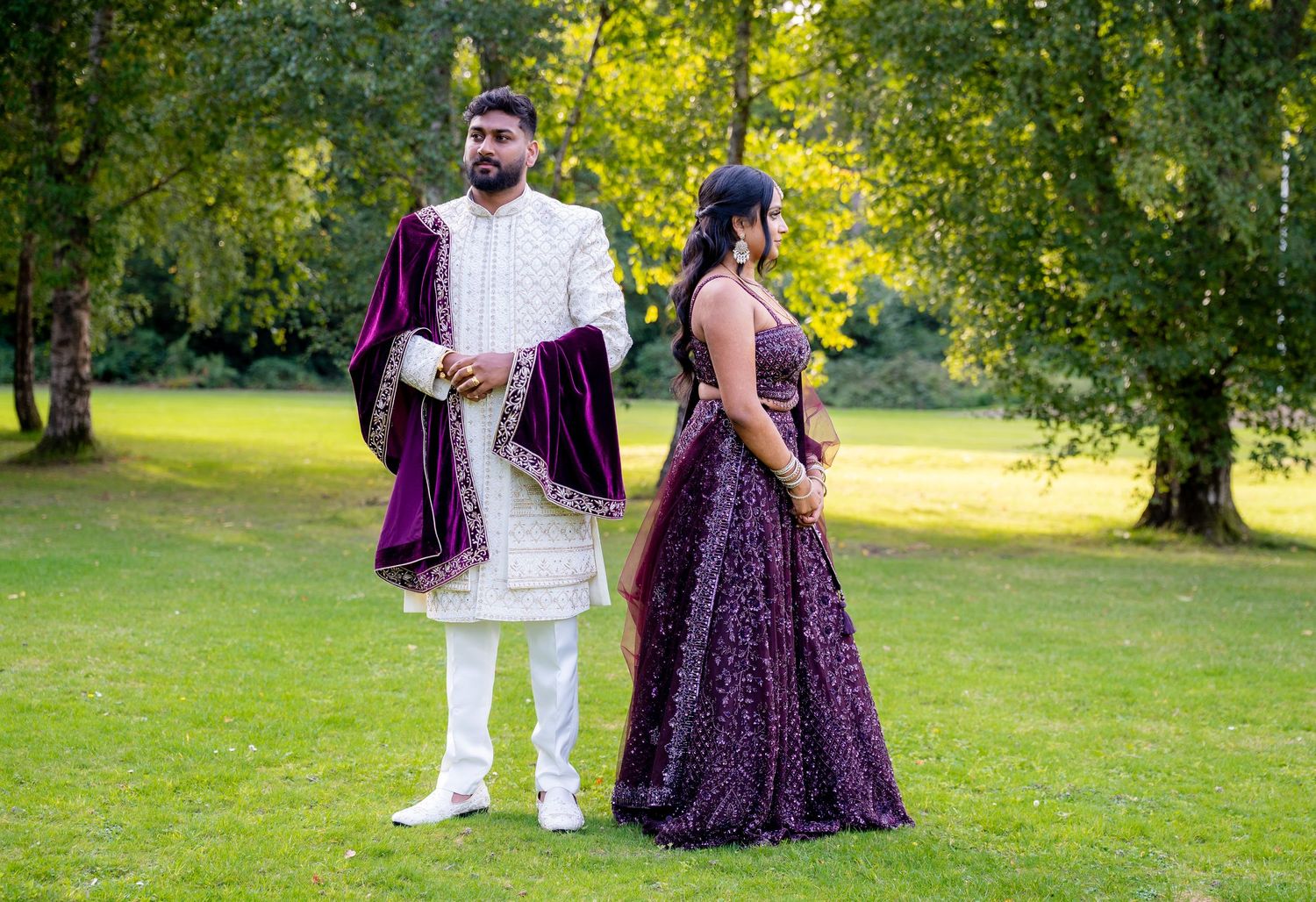 Couple in traditional Indian wedding attire, man in white sherwani with purple velvet shawl, woman in purple lehenga, outdoors.