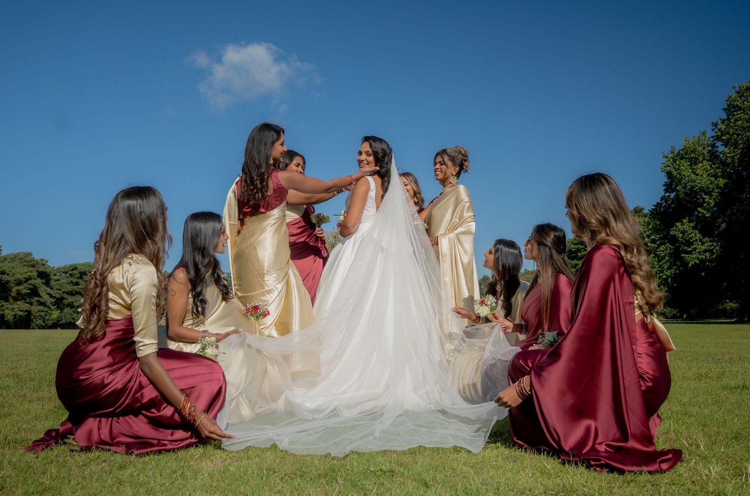 Bride in white gown surrounded by bridesmaids in burgundy and gold dresses on green lawn under blue sky.