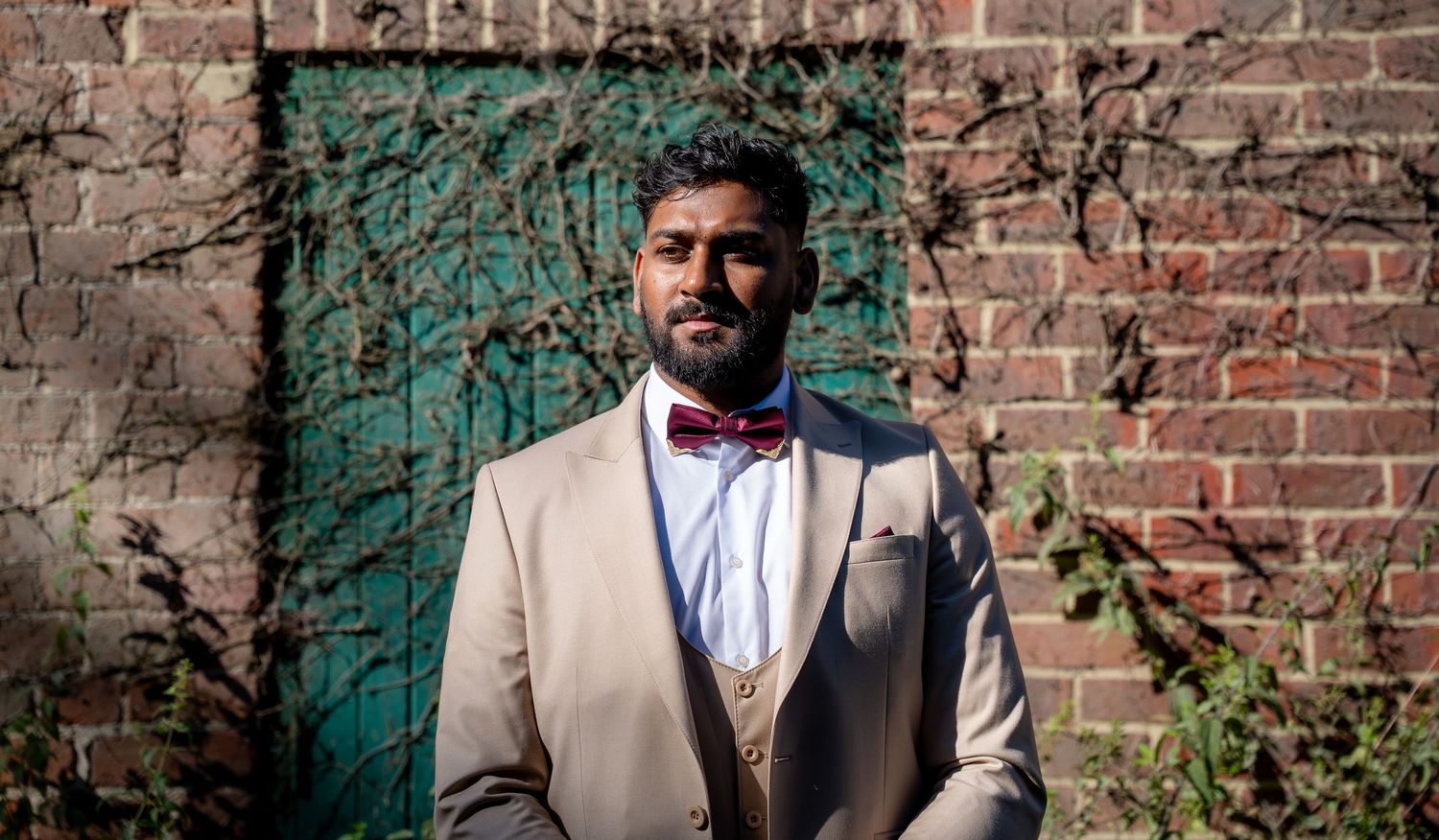Stylish man in beige suit with red bow tie posing against brick wall with green door.