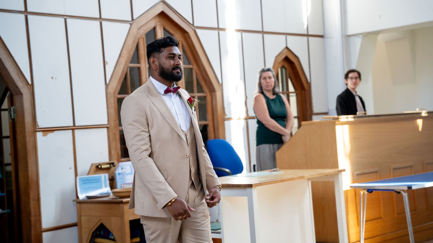 Groom in beige suit with burgundy bow tie stands at church altar during wedding ceremony.