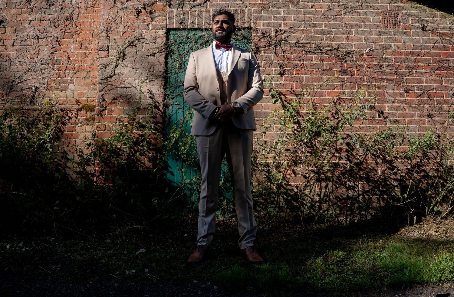 Groom in tan suit poses against a weathered brick wall with green door.
