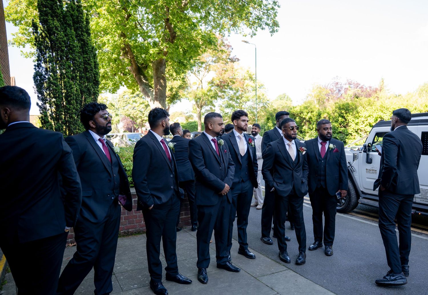 Groomsmen in dark suits with burgundy ties standing outside on sunny day before wedding ceremony.