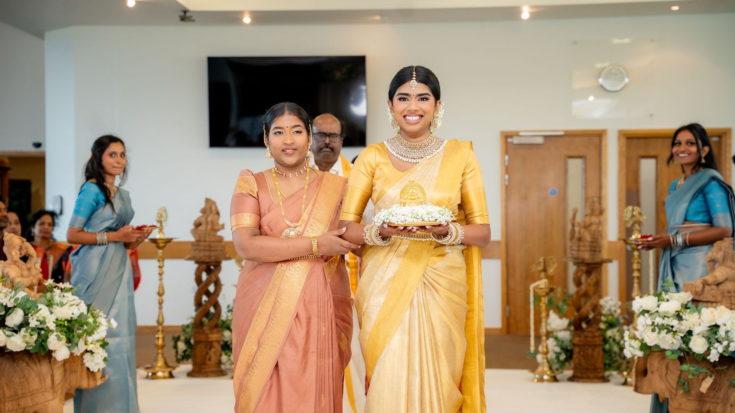 Two women in traditional Indian silk sarees smile while holding a decorative plate during a South Indian wedding ceremony.
