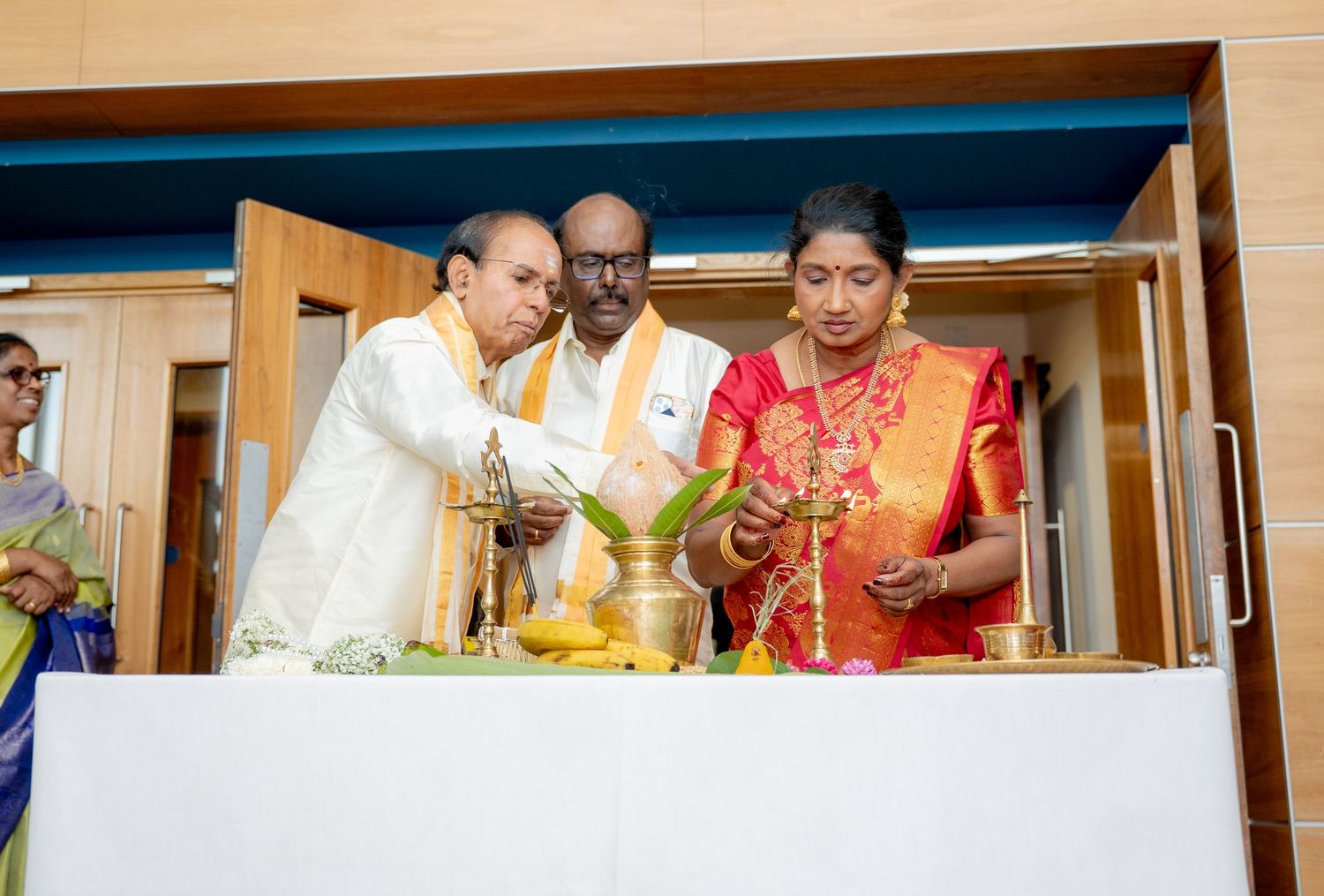 Two men and a woman perform a traditional Hindu puja ceremony with brass lamps and offerings on a white table.