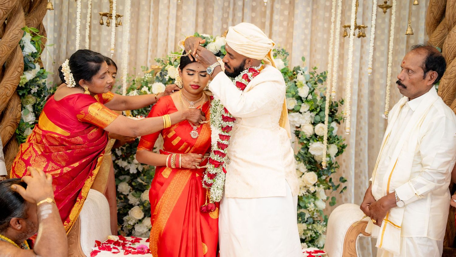 South Indian wedding garland exchange ceremony with bride in red saree and groom in white sherwani with family.