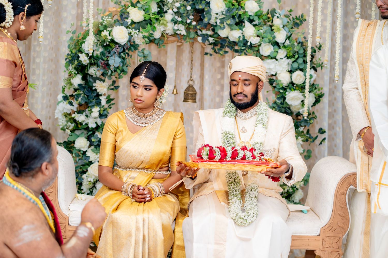 South Asian couple in traditional wedding attire seated at a floral mandap during a Hindu marriage ceremony.