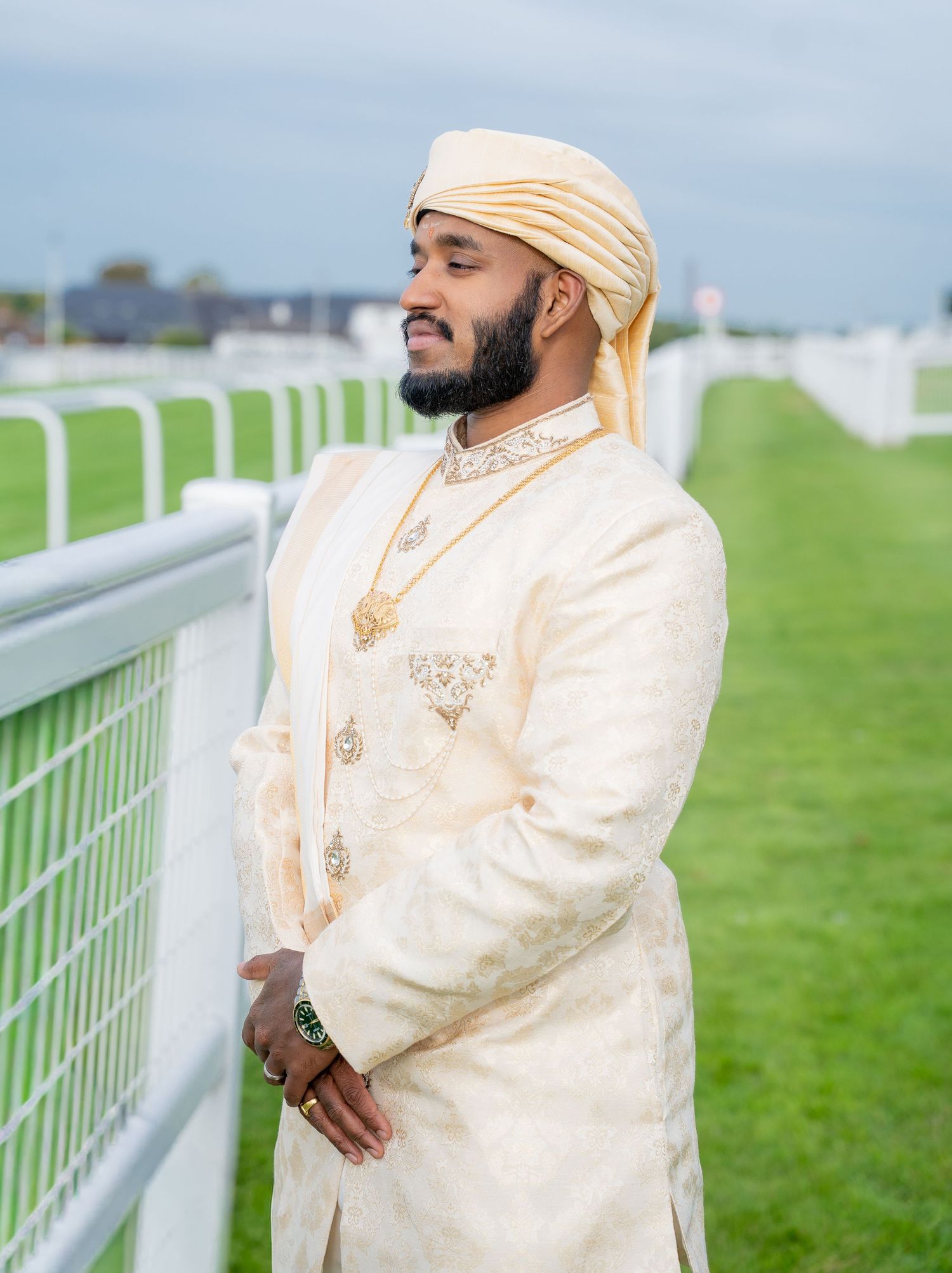 A man in cream sherwani and gold turban stands by a white fence on a green racecourse, smiling gracefully.