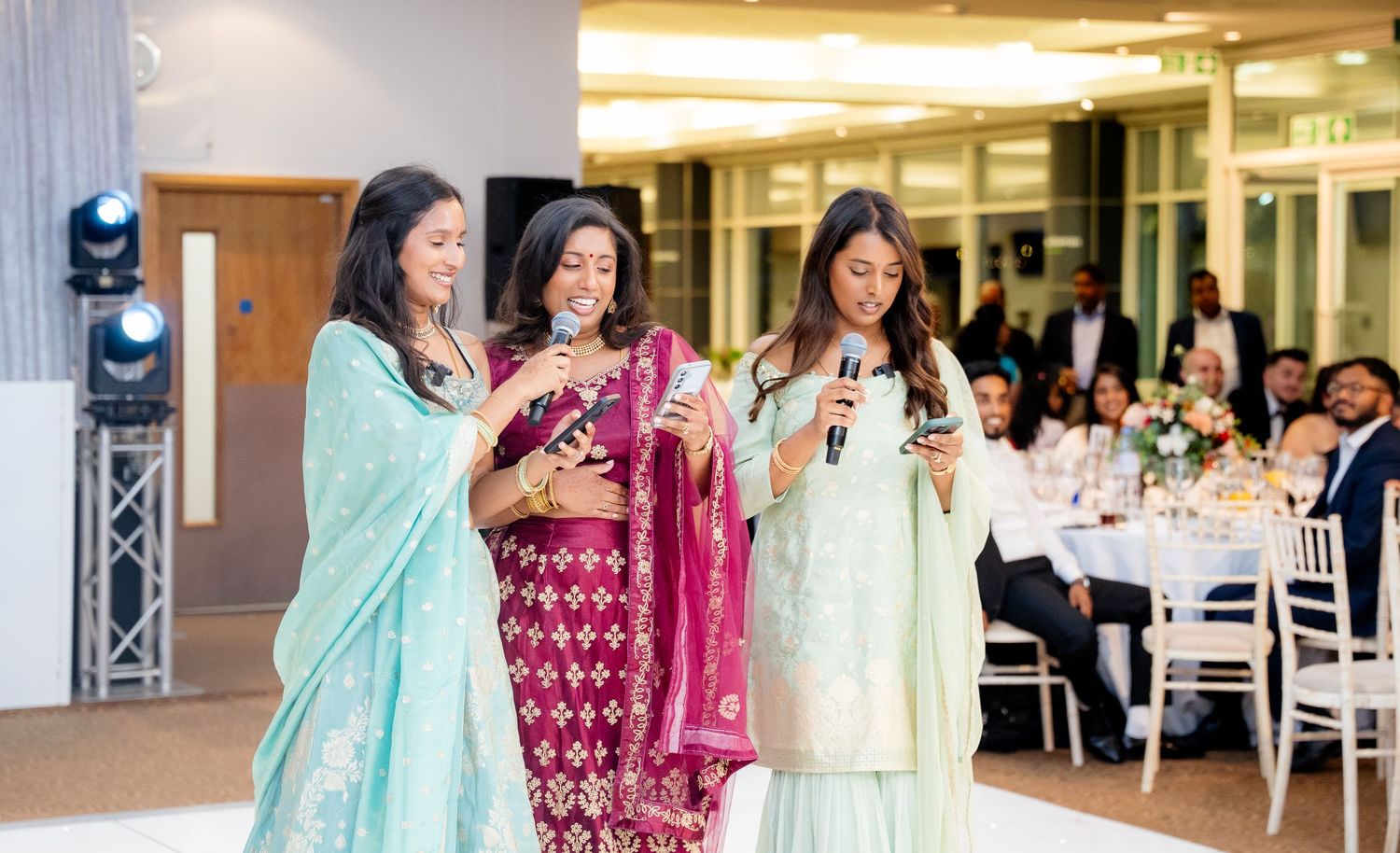 Three women in mint and pink Indian suits perform with microphones at an elegant wedding reception.
