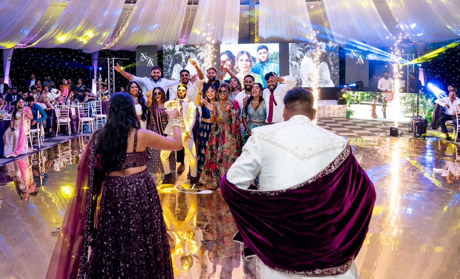 Colorful Indian wedding reception celebration with guests dancing on a golden dance floor under a decorated tent.