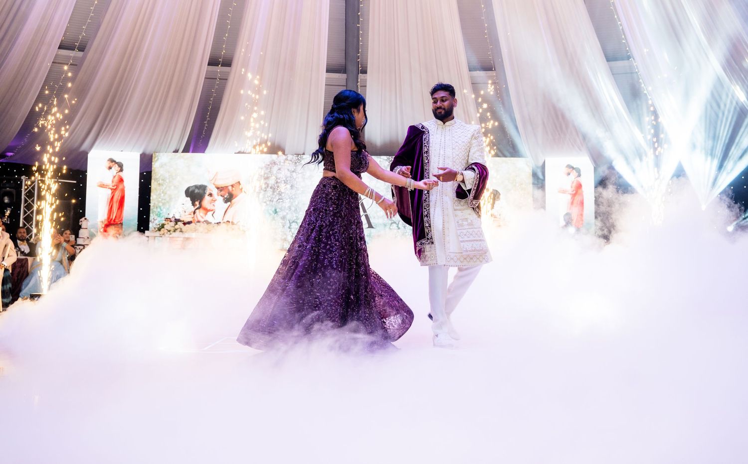 Bride in purple lehenga and groom in white sherwani share first dance surrounded by smoke and sparks.