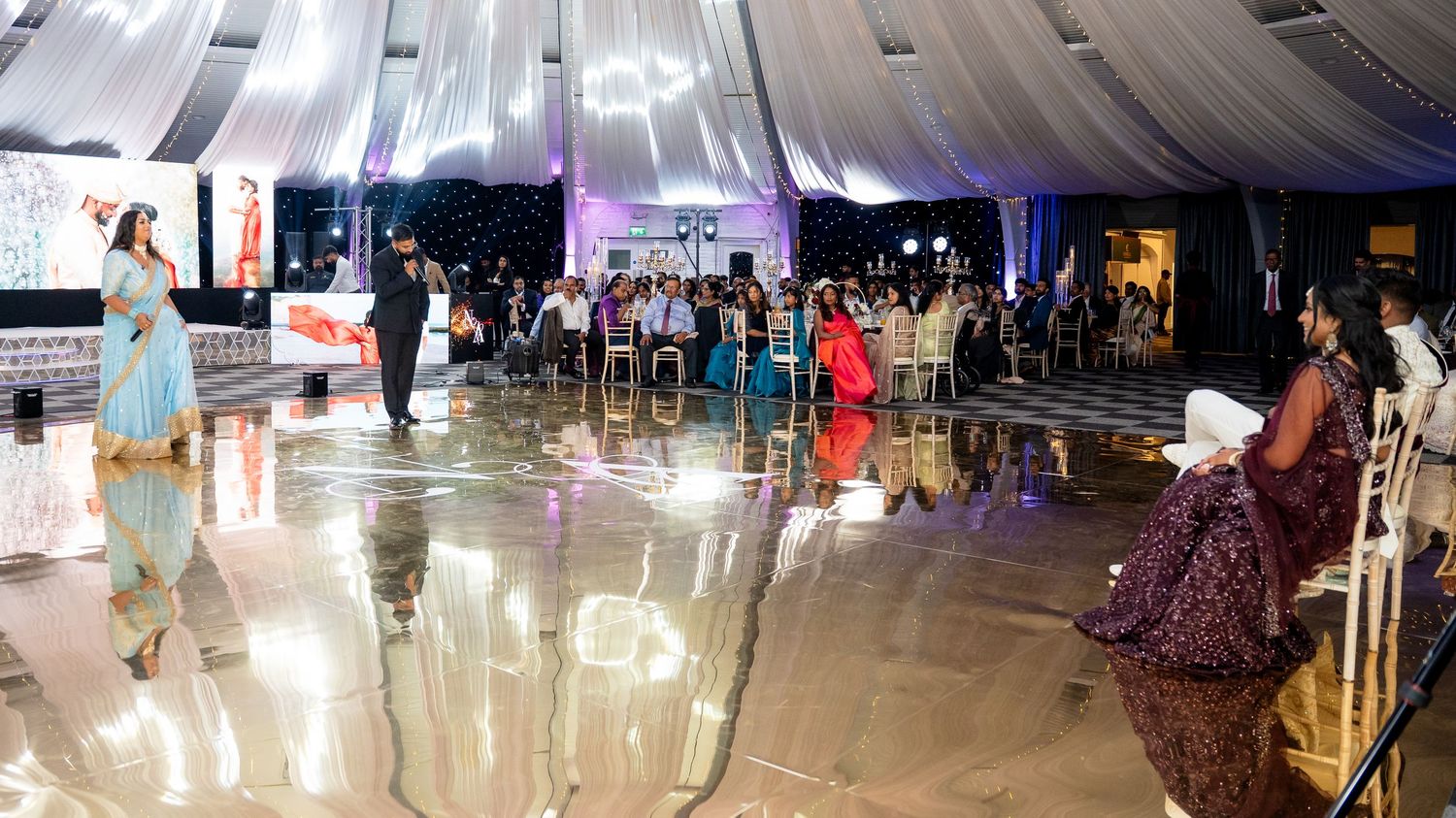 Guests dancing on a reflective floor at an Indian wedding reception in a decorated tent.