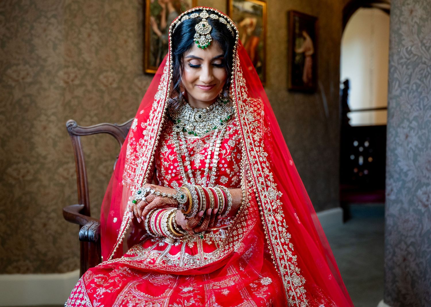 Indian bride in red lehenga with gold bangles and mehndi sitting gracefully on chair.