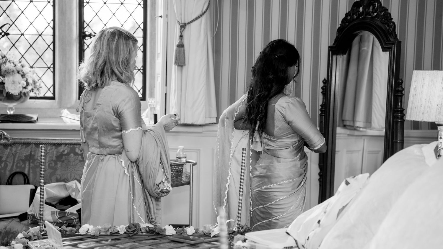 Two women in formal gowns prepare bridal dress in elegant dressing room mirror.