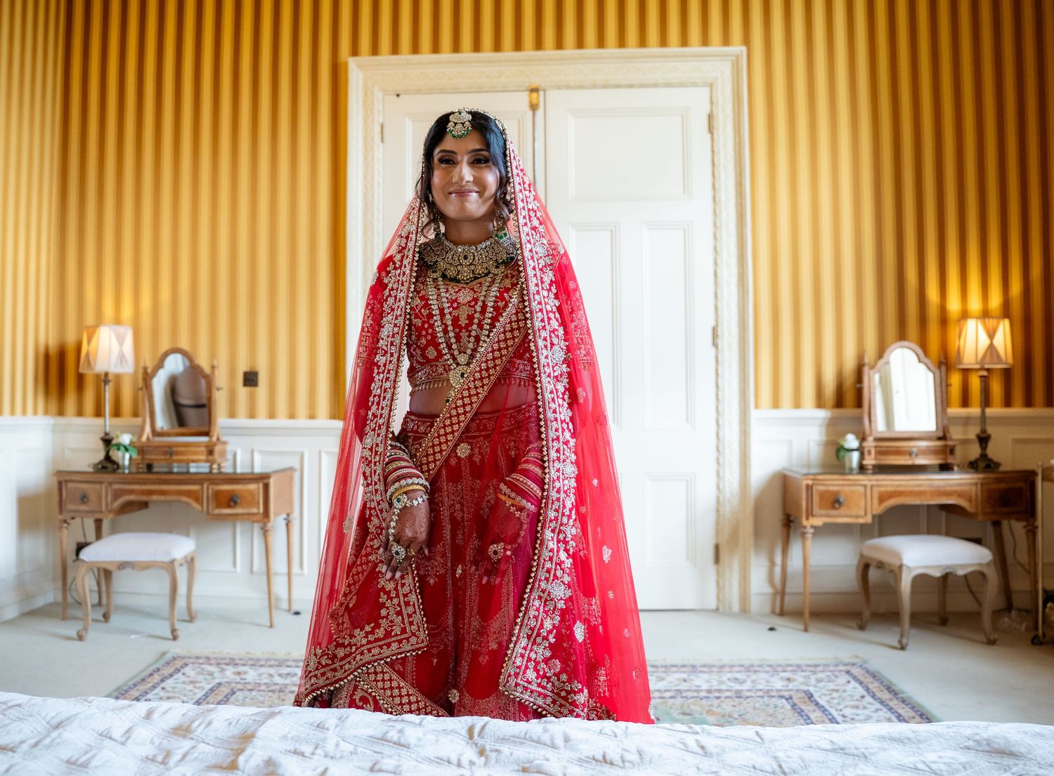 Indian bride in stunning red embroidered lehenga with gold jewelry standing in elegant bedroom.