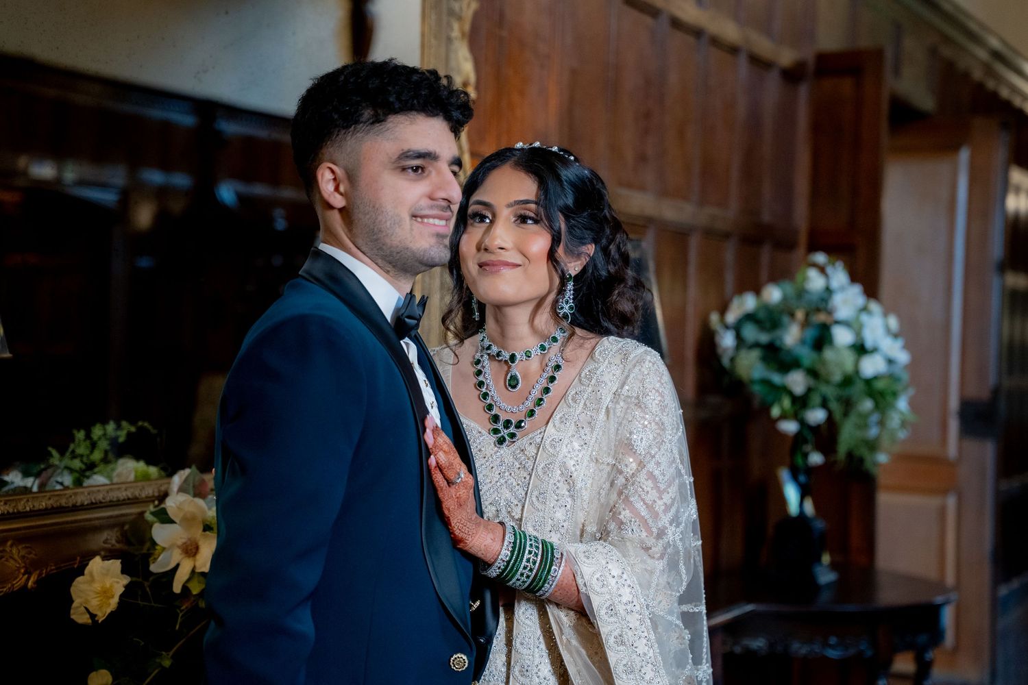 South Asian bride and groom in traditional wedding attire share an intimate moment in ornate wood-paneled hall.