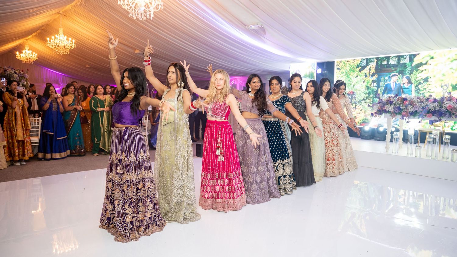 Women in colorful lehengas dancing together at an elegant Indian wedding reception under a white tent.
