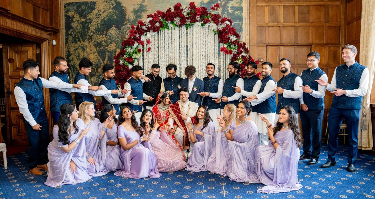 Wedding party poses under floral arch with bridesmaids in lavender and groomsmen in black sherwanis.