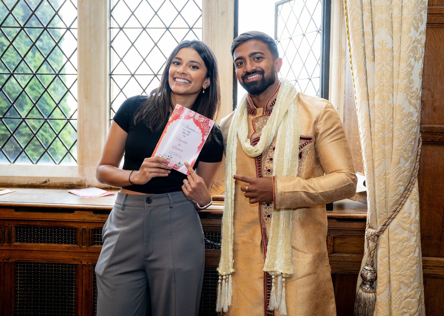 Young couple posing with wedding invitation card at an Indian wedding venue.