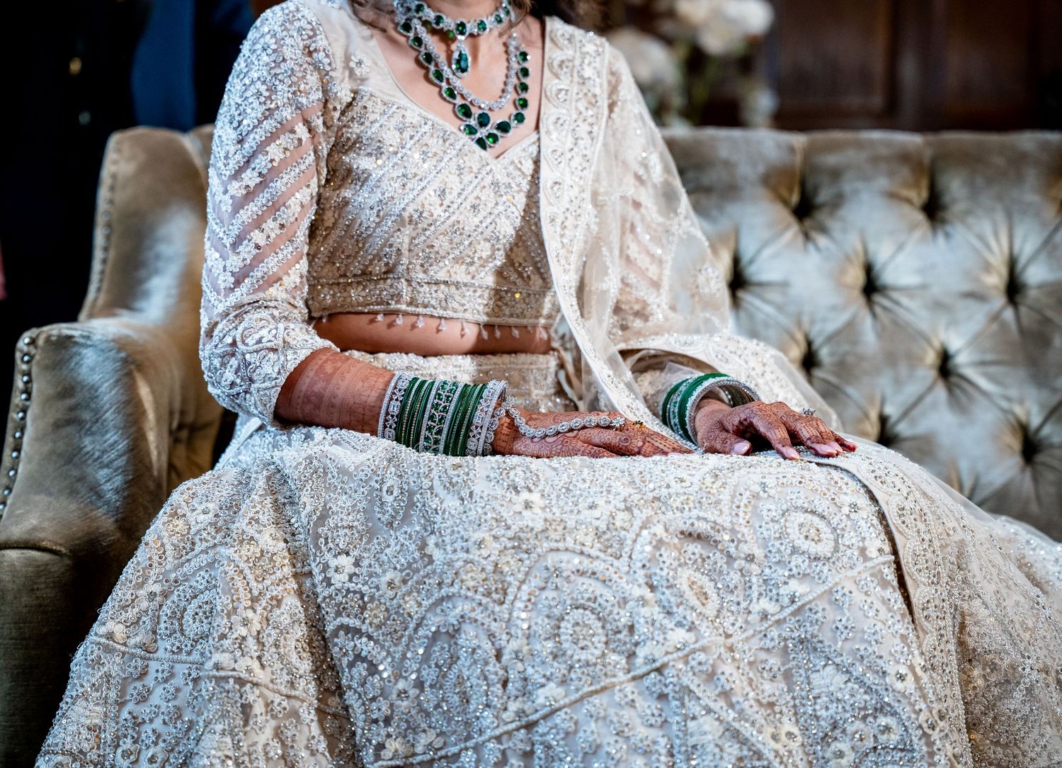 Indian bride in ivory embroidered lehenga with green bangles and henna hands seated on tufted sofa.