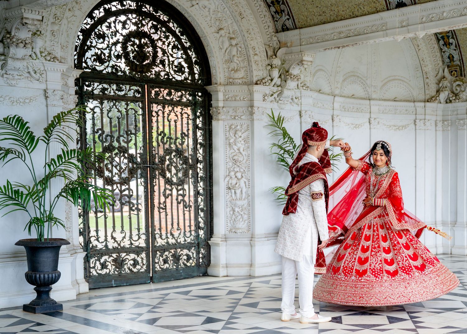 Indian bride and groom in traditional red and white wedding attire pose by ornate iron gates at a marble palace.