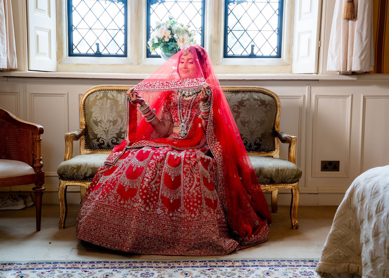 Indian bride in red embroidered lehenga seated on ornate sofa near Gothic arched windows.
