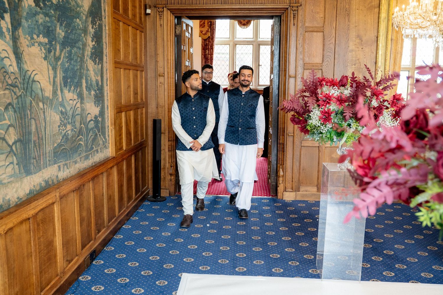 Two men in traditional shalwar kameez walk through elegant hallway decorated with floral arrangement.