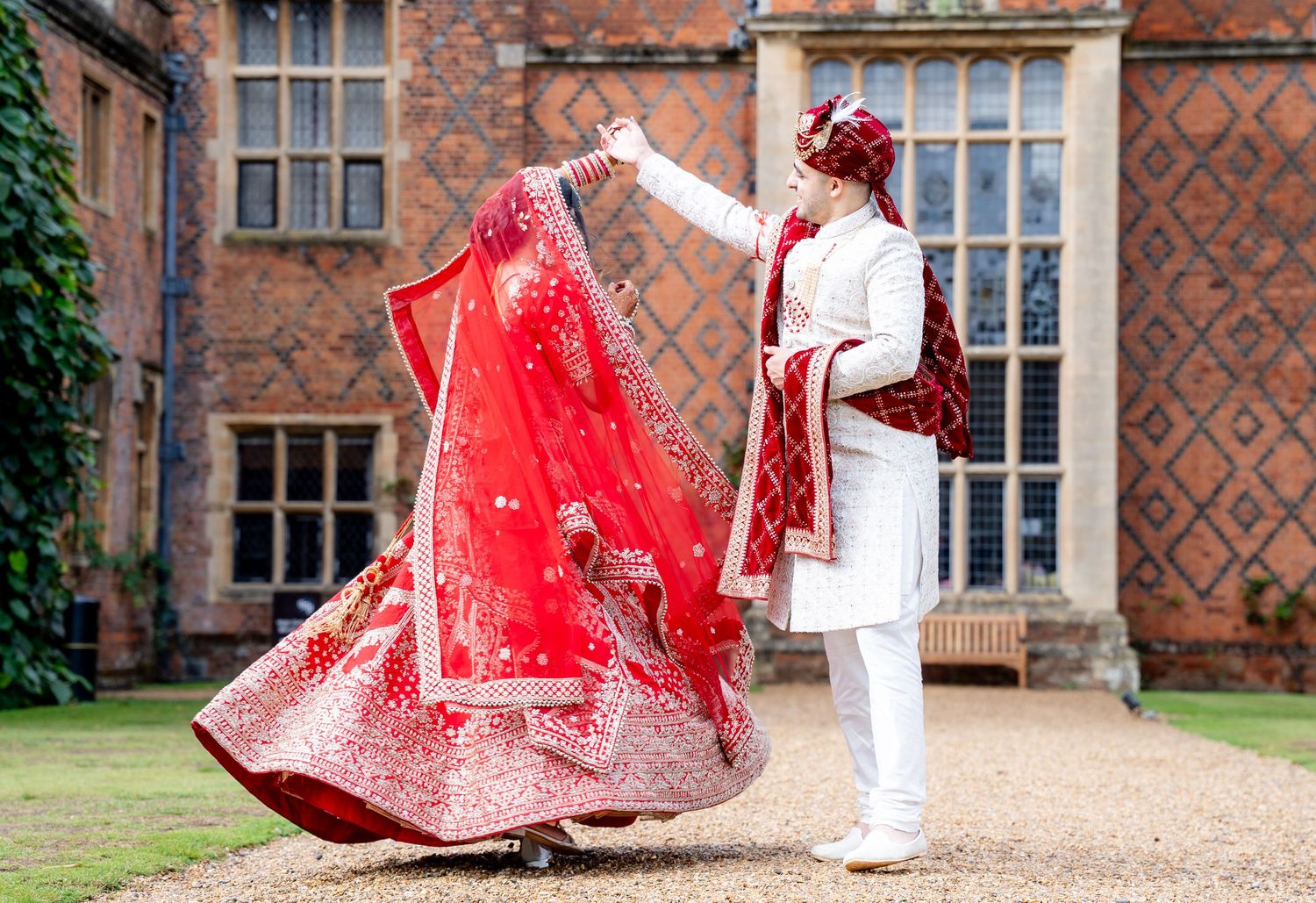 Indian bride in red lehenga twirling with groom in white sherwani at elegant brick manor wedding venue.