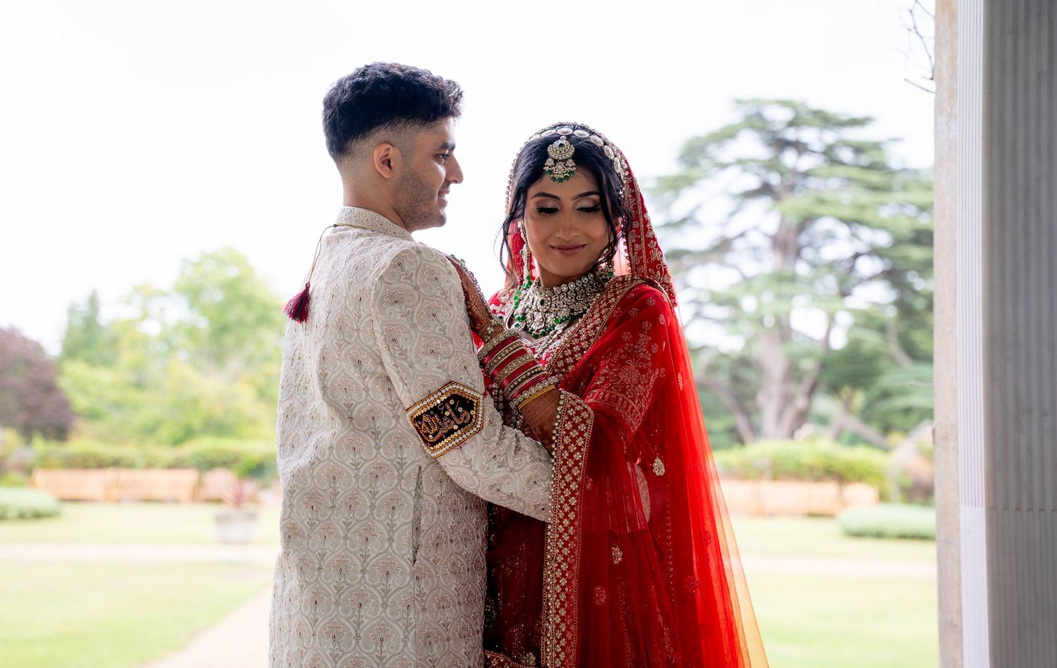 South Asian bride in red lehenga and groom in cream sherwani share a tender moment outdoors at a wedding.