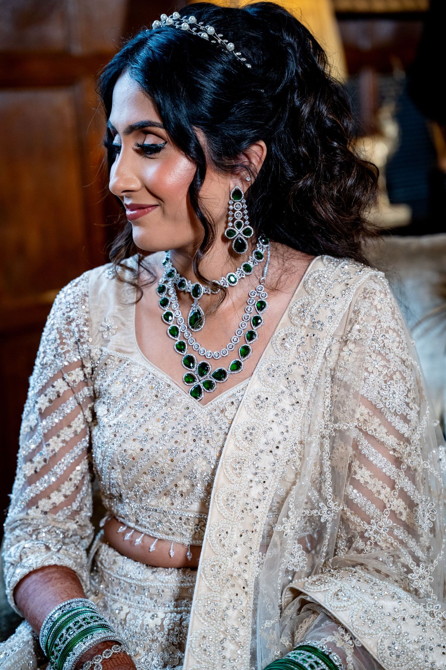 Indian bride wearing emerald and diamond jewelry with white embroidered lehenga and floral hair accessory.