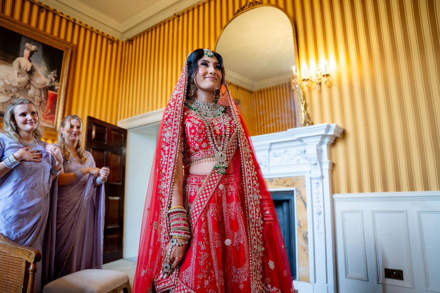 Indian bride in red lehenga smiling joyfully surrounded by guests in ornate gold-wallpapered wedding room.