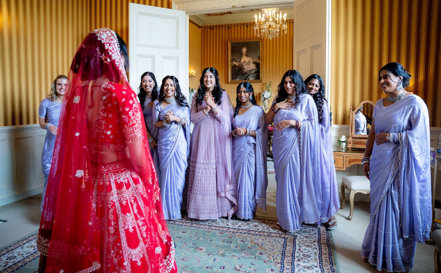 Indian bride in red lehenga revealed to bridesmaids in lavender sarees.