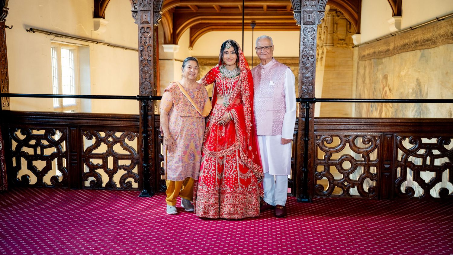 Indian bride in red lehenga poses with family on ornate balcony during traditional wedding ceremony.