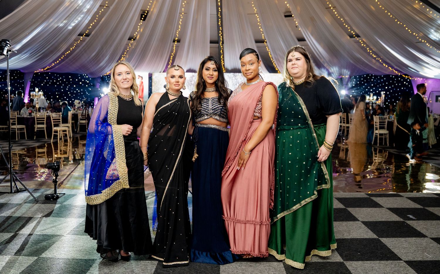 Five women in elegant Indian sarees pose together at a festive event with decorative lights in the background.