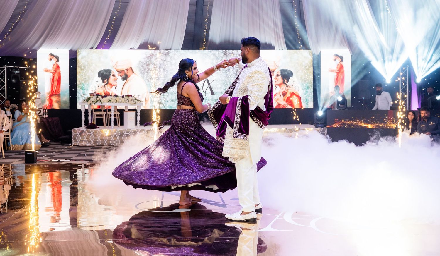 Couple dancing in purple attire on reflective floor with fog effects at an elegant indoor wedding reception.