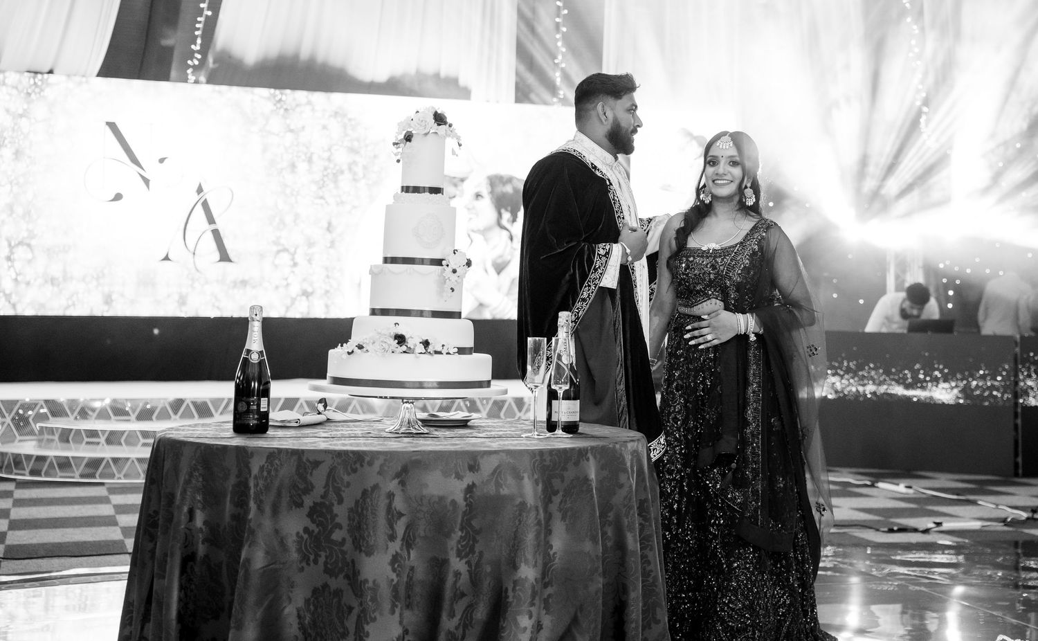 Black and white photo of bride and groom posing beside elegant tiered wedding cake with champagne bottles.