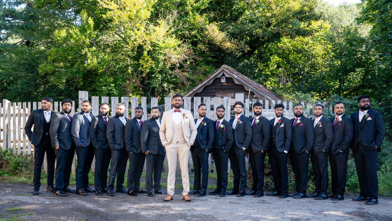 Groom in cream suit poses with groomsmen in black tuxedos in front of rustic wooden backdrop.