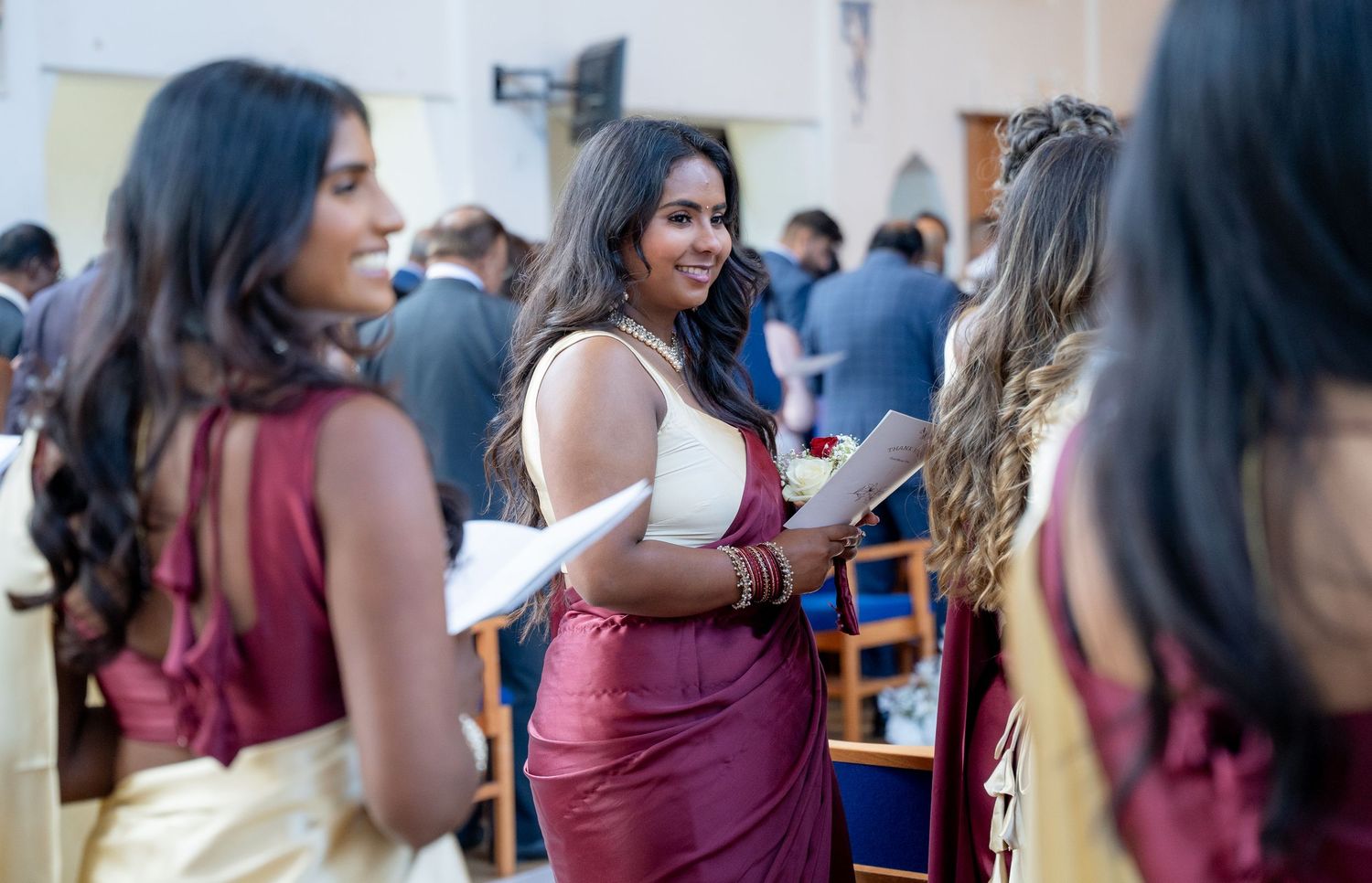 Bridesmaid in burgundy sari smiles warmly while chatting with guests at wedding reception.
